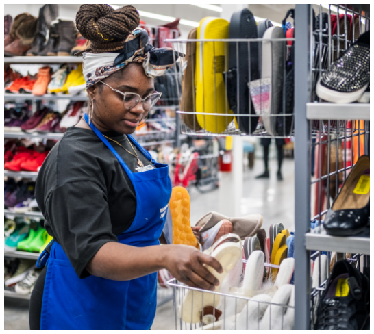 RWB Thrift employee in a blue vest organizing shelves of shoes.